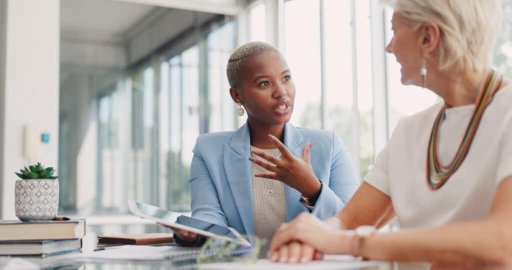 Two women at work discuss business while holding a tablet between them; demonstrating leadership and communication soft skills