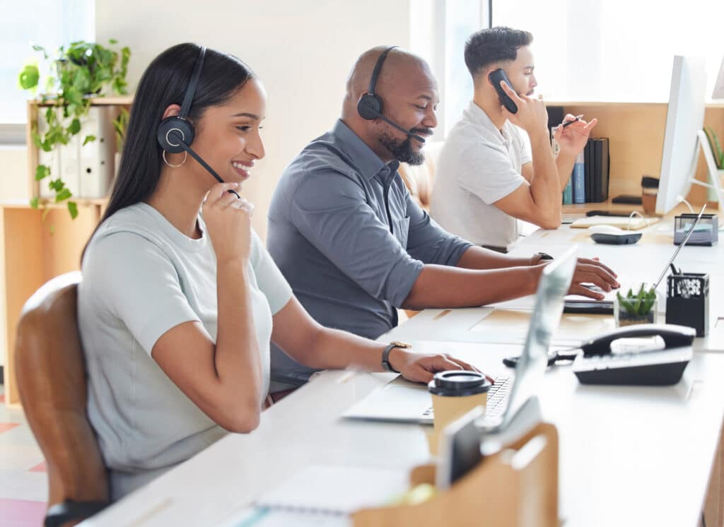 Three customer service representatives are taking phone calls while looking at computer screens in an office setting.