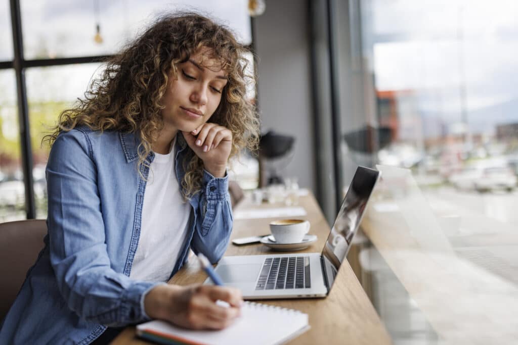 Woman sitting at a counter at a café in front of a laptop while writing in a notebook