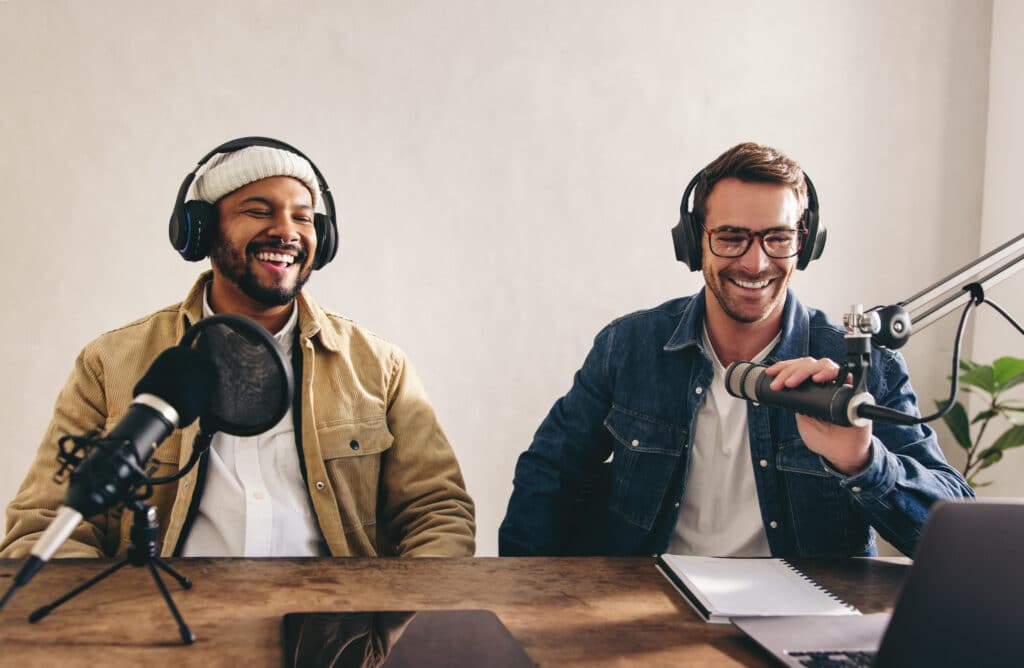 Two men laughing and sitting side by side in a recording studio wearing over-the-ear headphones in front of microphones 